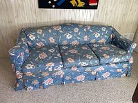 Full front view of the blue floral fabric sofa bed showing three loose seat cushions, floral upholstery, and pleated skirt base.
