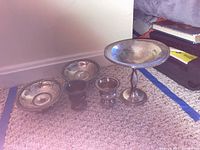 View of the five sterling silver pieces on carpet: two shallow bowls, one goblet, one pedestal dish, one lidded container.