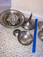 Eight sterling silver table accent pieces arranged on a carpeted floor, including bowls, trays, and a candlestick holder base with visible tarnish and patina, one piece appears damaged.
