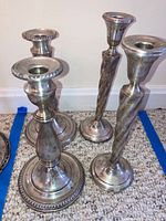 Four sterling silver weighted candlesticks on carpeted floor exhibiting two distinctive designs, with a mix of rounded and twisted stems and weighted bases.