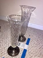 Two clear glass bud vases of different heights with weighted sterling silver bases on a carpeted floor against a baseboarded wall.