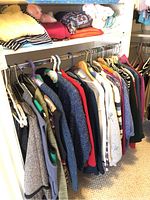 Various sweaters and shirts hanging on rack in closet, mostly in small and medium sizes, folded clothing and a doll on shelf above