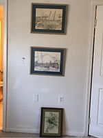 Three framed art prints hung and leaning against a wall in a room. Two show sailboats in black and white, one shows birds on a branch with green foliage.