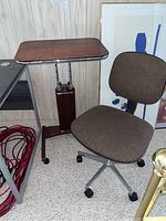 Wide shot showing the writing table next to a brown upholstered chair, both placed against a wooden paneled wall on carpet.
