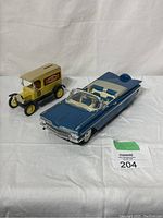 Blue and white 1959 Chevrolet Impala die-cast model and yellow 1917 Ford Model T van piggy bank on white cloth background with tag 204
