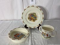 Front view of the Bunnykins plate, bowl, and two-handled mug arranged on stands and flat surface
