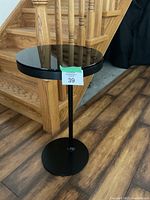 Full view of round Ikea end table with metal base and column, dark glass top, and wood accents beneath glass.