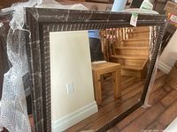 Photo of large rectangular mirror with dark distressed wood frame leaning against wall in room with wooden furniture and staircase.