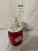 Front view of ceramic tea canister showing red barrel body, TEA label, and bust lid with glasses.
