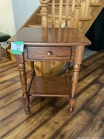 Front view of a solid oak end table showing a single drawer and a bottom shelf with turned legs.
