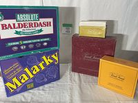 Five vintage board games displayed including Absolute Balderdash, Malarky, and three editions of Trivial Pursuit in varying box sizes and colors on a white fabric surface.