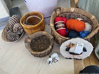 Photo showing five woven baskets of different sizes, six skeins of brightly colored yarn, two small ceramic figurines of a ewe and lamb, and several large pinecones on and near the floor.