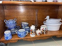 Photo of assorted blue and white Japanese china cups, saucers, a sugar bowl, and creamer with intricate floral and scenic designs, a clear glass vase, small commemorative English china pieces with flag designs, and a white ceramic duck-shaped casserole dish with lid on a wooden shelf.