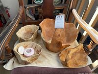 Four hand-carved wooden items displayed on a chair; three bowls and one basket with natural wood grain and cracks visible.