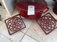 Large red enameled cast iron Dutch oven with lid and two coated trivets on tiled surface.