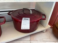Photo of red Martha Stewart enameled cast iron Dutch oven on shelf showing side handles, lid with stainless steel handle, and attached lot tag.