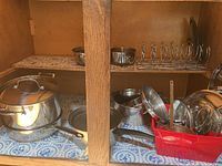Photo of a cabinet shelf with various Earth Chef stainless steel pots, pans, lids, a metal colander, and a metal toast rack.