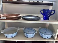 Photo showing 8 copper-colored round chargers, 1 dark round plate, 3 white ceramic casserole dishes with blue floral patterns, and blue glass vase on shelves.