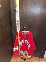Denver Pioneers red hockey jersey hanging with wooden left-handed hockey stick and two pucks on the floor.