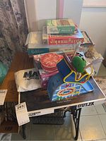 Photo shows a variety of board games and puzzles stacked on a table, including Wild Thing marble game, multiple puzzle boxes, Yahtzee, and cloth bag labeled 'African'.