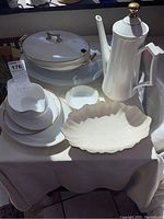 Overall view of eight porcelain serving items with gold trim on a white tablecloth including plates, a covered dish, a scalloped bowl, and a coffee pot.