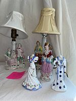 Two small lamps with fabric shades and five porcelain figurines including seated couple and standing women in traditional dress.