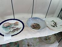 Three porcelain plates displayed on a shelf, featuring different painted decorations including fish, birds, water lilies, and embossed rims.