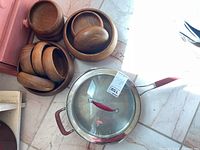 Two sets of nested teak wood salad bowls arranged on a surface beside the metallic frying pan with glass lid and red handles.