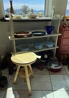Painted wooden shelf with multiple shelves displaying various bowls, dishes, and decor, beside small wooden swivel stool on tile floor