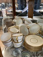 Display showing multiple ivory tea cups and gold-trimmed saucers, some stacked and some upright, with various gold edge patterns visible.
