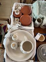 White fabric-lined box with stacked reddish-brown terracotta plates and small cups on a chair next to ceramic jugs and bowls.