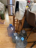 Overview photo of the ceramic water cooler on a wooden stand with fabric cover and four large plastic water jugs on floor.