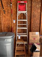 Two ladders in garage setting, one metal folding ladder with red top and one wooden folding ladder below it.