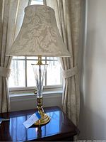 Single matching lamp on dark wood side table with beige patterned shade, positioned near window with patterned curtains.