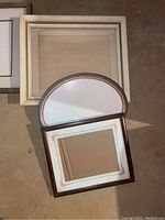 Photo of three frames stacked lying on floor, shows cream-white rectangular frames and oval bronze frame