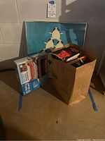 Overall view of the lot showing a large paper bag containing multiple VHS tapes, a boxed spice rack organizer beside it, and a framed penguin print behind.