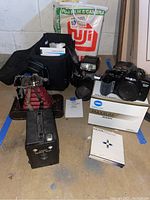 Photo of all cameras and accessories including Minolta Maxxum 3200i and 3000i cameras, Hoya filter box, Target Brownie Six-20 camera, Kodak folding camera, camera bag and flash unit on floor.