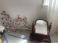Dresser top mirror with dark wooden frame and turned finials on a base, placed beside pink flower metal wall hanging on the floor near a window.