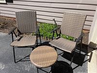 Two outdoor folding chairs and one small folding end table displayed outdoors on pavement against house siding. Chairs and table have woven synthetic rattan-style surfaces and black metal frames.