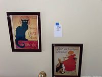 Photo showing two framed French prints hanging on a wall, one with a black cat and the other with a girl in red dress and cats.