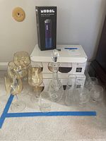 Two boxes of Libbey 20 oz clear wine glasses (12 total), assorted tinted and clear glassware including stemless champagne glasses, red and white wine glasses, small glasses, a glass decanter with stopper, a glass carafe, and a boxed Modal battery-operated wine opener all arranged on a white table.