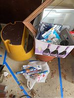 Yellow rolling step stool next to terra cotta plastic flower pot and box with assorted household items including light bulbs, frame, brush, and switch plate covers.
