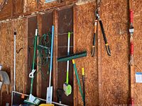 Photo showing an array of long-handled tools hanging against an unfinished wooden wall in a garage. Visible tools include a green push broom, mops, long handled brushes, and extension cords.