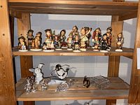Two wooden shelves with 14 Hummel figurines on the top shelf and assorted glass and porcelain figurines on the bottom shelf.