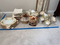 Overall view of stacked Noritake china pieces including plates, saucers, bowls, and cups arranged on carpet.