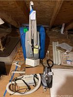 Photo showing the upright vacuum, canister vacuum with hose and strap, small travel iron, clothes steamer, and large blue suitcase stored in attic space on plywood floor.