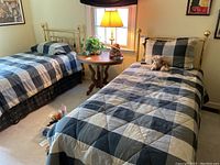 View of two twin beds with brass headboards, dressed in black and gray patchwork bedding around a round wood nightstand with a lamp.