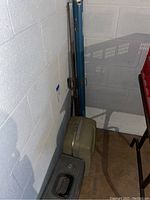 Photo showing vintage slide projector case and blue metal folding screen case leaning against basement wall.