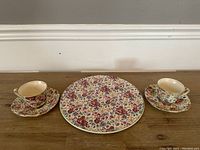 Full view of two Royal Winton tea cups and saucers flanking a Royal Tudor floral chintz platter on wooden surface against plain wall