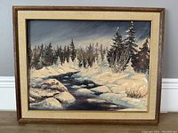Framed oil painting showing snowy river scene with pine trees, wooden frame with beige matting visible.